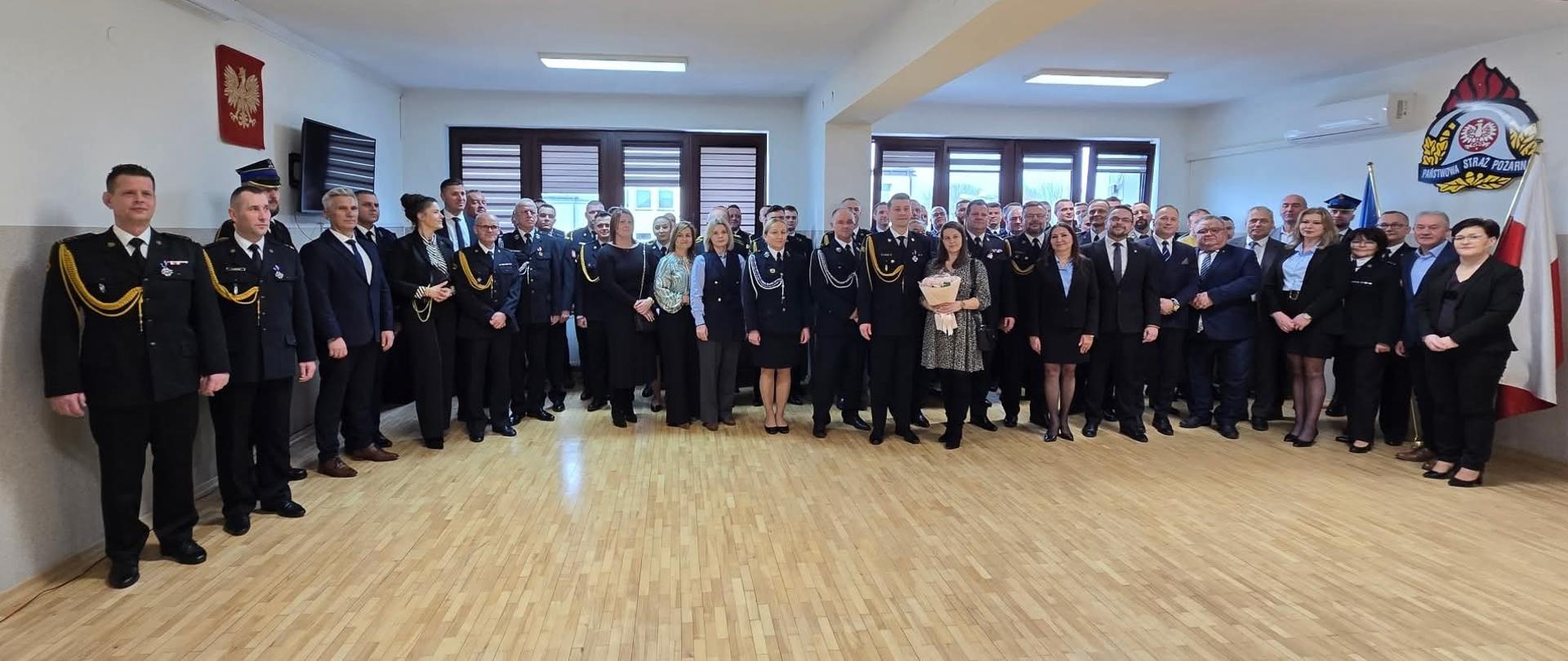 Zdjęcie grupowe wszystkich uczestników wydarzenia. Zdjęcie zrobione na jasnej sali z białymi ścianami i drewnianą podłogą. 