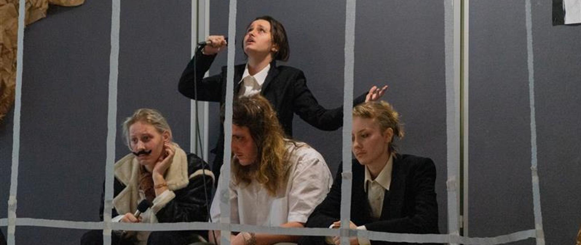 Scena przedstawia więzienie, na pierwszym planie widoczna krata. Trzy osoby siedzą, jedna stoi a nimi i mówi do mikrofonu.