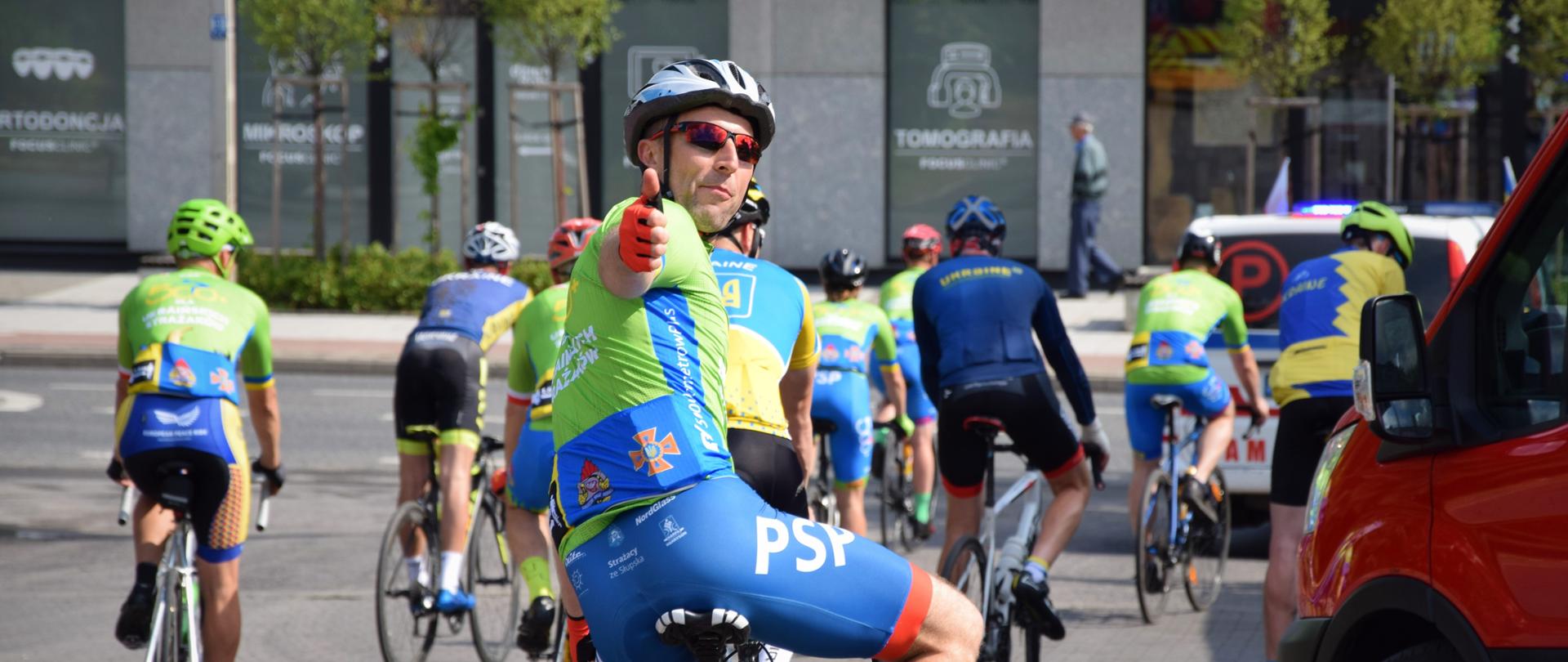 Zdjęcie zrobione z tyłu kolarzy, kolarze odjeżdżają z tut. Komendy, po kolei wjeżdżają na asfalt celem ruszenia w dalszą trasę rowerową, jeden z kolarzy odwrócił się do tyłu (przodem do aparatu) i pokazuje kciuki w górę (tzw. like)