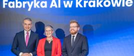 Na tle wielkiego ekranu z napisem "Fabryka AI w Krakowie" na niebieskim tle. stoją trzy osoby, wiceminister Mrówczyńska w czerwonej marynarce pośrodku między dwoma mężczyznami w czarnych garniturach.