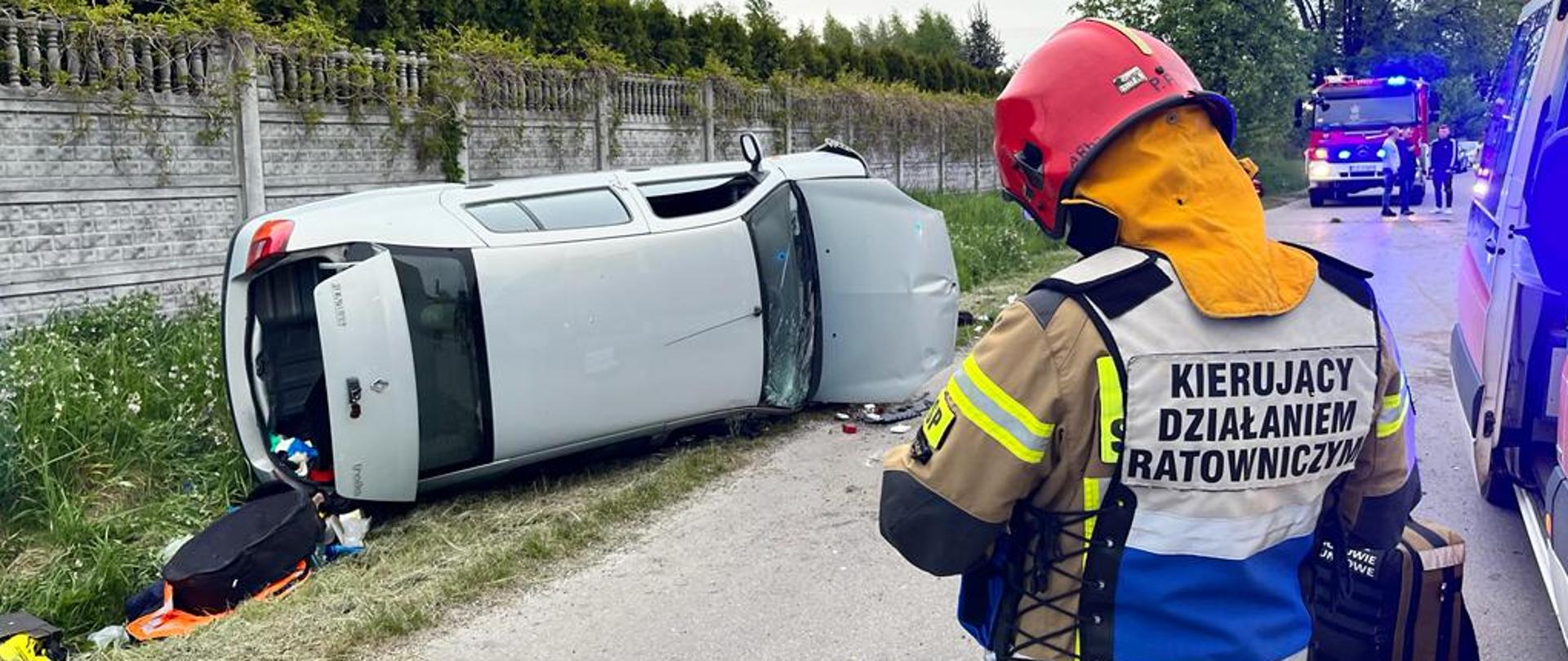 Na zdjęciu widoczny samochód osobowy na prawym boku w rowie. Na pierwszym planie widać strażaka z kamizelką „Kierujący działaniem ratowniczym”, który sporządza niezbędną dokumentacje na miejscu zdarzenia. 