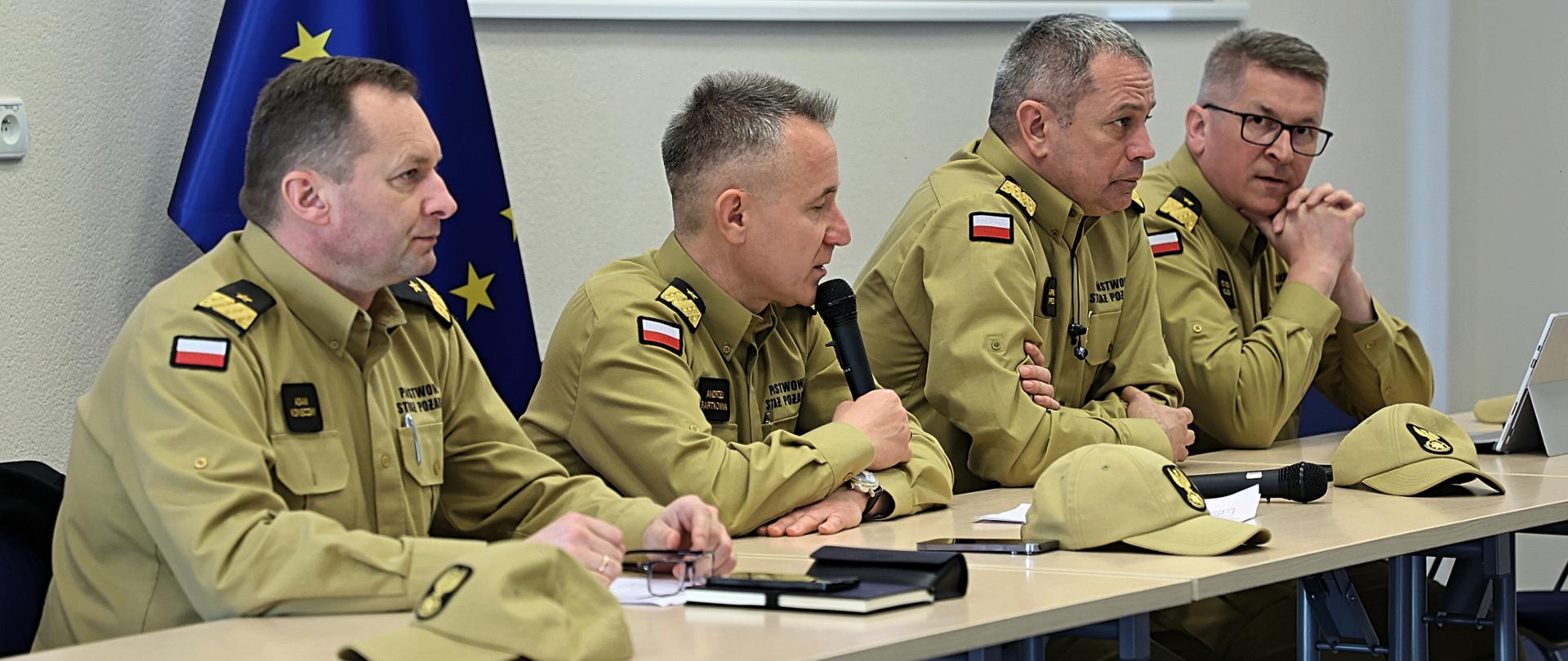 Na zdjęciu widać od lewej zastępcę komendanta głównego PSP nadbryg. Adama Koniecznego, komendanta głównego PSP nadbryg. Andrzeja Bartkowiaka, zastępców komendanta głównego nadbryg. Arkadiusza Przybyłę i nadbryg. Krzysztofa Hejduka którzy siedzą przy stoliku przemawiając do zebranej kadry. w tle flaga Unii Europejskiej oraz prezentacja.