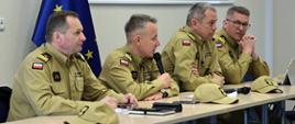 Na zdjęciu widać od lewej zastępcę komendanta głównego PSP nadbryg. Adama Koniecznego, komendanta głównego PSP nadbryg. Andrzeja Bartkowiaka, zastępców komendanta głównego nadbryg. Arkadiusza Przybyłę i nadbryg. Krzysztofa Hejduka którzy siedzą przy stoliku przemawiając do zebranej kadry. w tle flaga Unii Europejskiej oraz prezentacja.