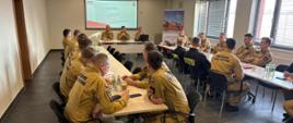 Grupa strażaków w mundurach siedzi przy stołach w sali konferencyjnej podczas spotkania szkoleniowego; z przodu kilku prowadzących omawia prezentację wyświetlaną na ekranie.