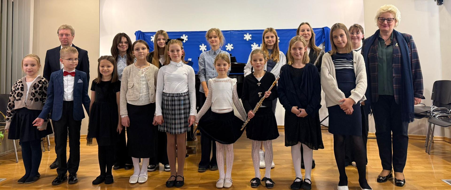 Na zdjęciu odświętnie ubrani uczniowie, którzy są ustawieni w dwóch rzędach. Zdjęcie zostało wykonane podczas koncertu uczniów klasy fletu Pani Katarzyny Karczewskiej oraz fortepianu Pana Zbigniewa Kąkola. Po lewej stronie obrazka w drugim rzędzie mężczyzna blondyn w czarnej marynarce, Po prawej stronie kobieta blondynka w okularach, czarnych spodniach, kraciastej marynarce z niebieskim szalikiem. W tle niebieski materiał z białym śnieżynkami. Podłoga wykonana z brązowego parkietu.