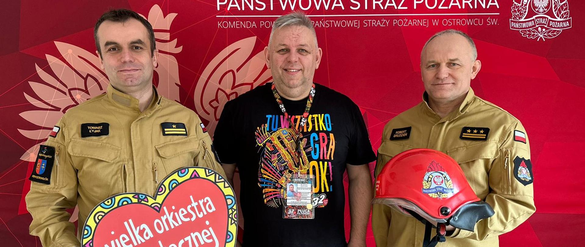 W siedzibie KP PSP w Ostrowcu Św. stoi trzech mężczyzn i pozuje do zdjęcia. Dwóch strażaków - komendantów powiatowych PSP w Ostrowcu Św. oraz szef sztabu WOŚP Ostrowiec Św. Jeden ze strażaków trzyma w rękach serduszko WOŚP, a drugi hełm strażacki przekazany do licytacji