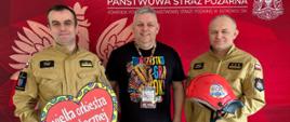 W siedzibie KP PSP w Ostrowcu Św. stoi trzech mężczyzn i pozuje do zdjęcia. Dwóch strażaków - komendantów powiatowych PSP w Ostrowcu Św. oraz szef sztabu WOŚP Ostrowiec Św. Jeden ze strażaków trzyma w rękach serduszko WOŚP, a drugi hełm strażacki przekazany do licytacji