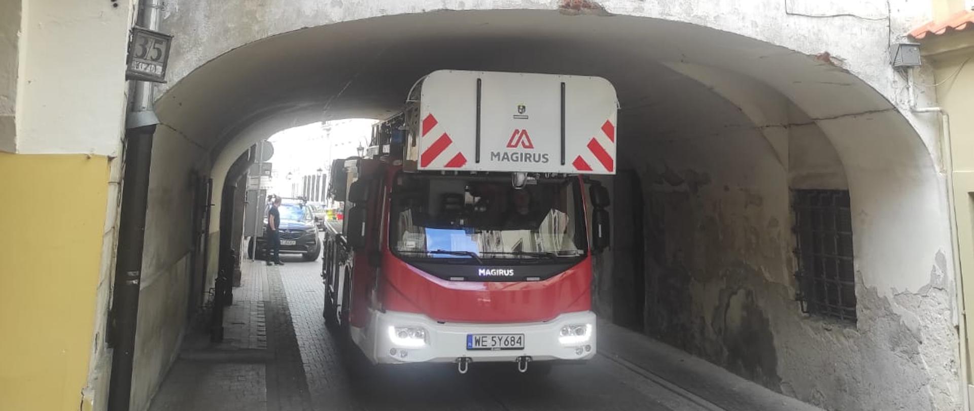 Wóz strażacki z drabiną mechaniczną przejeżdżający przez przejazd o niskim sklepieniu pomiędzy budynkami na Warszawskiej Starówce. Na zdjęciu widać drabinę pożarniczą koloru czerwonego z elementami bieli, która przejeżdża pod obniżeniem przy ul. Koziej.