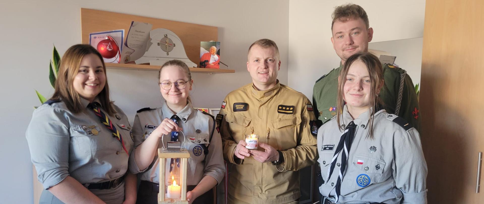 Zdjęcie przedstawia strażaka i harcerzy, którzy przekazują zapaloną święczkę.