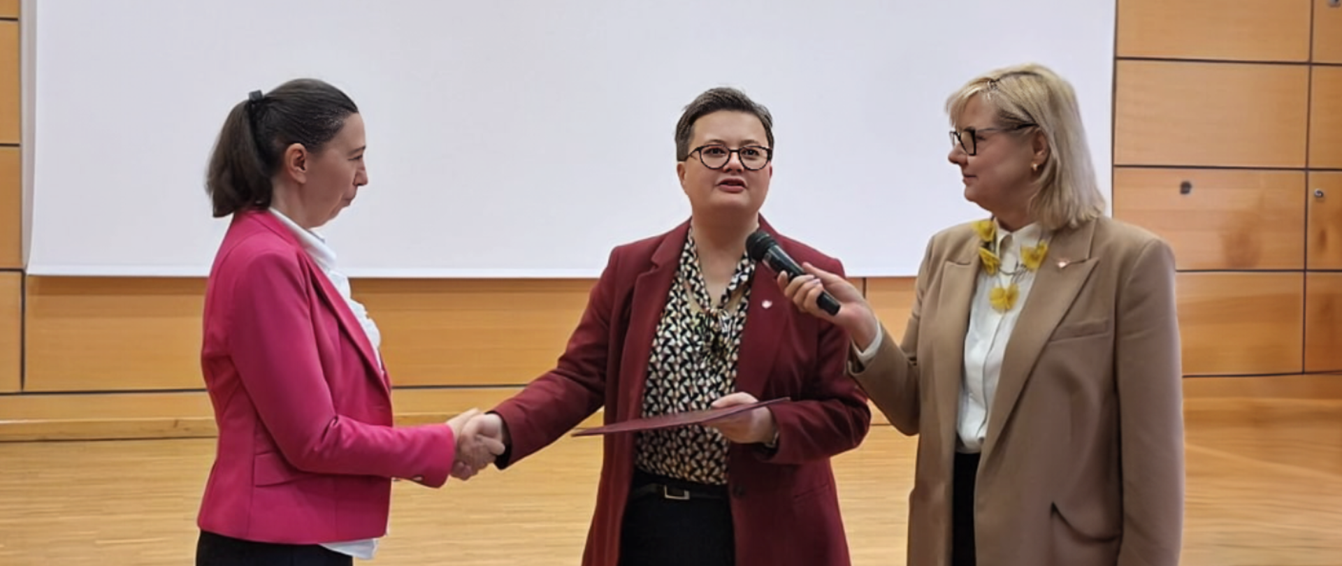 Wiceminister Lubnauer podaje rękę nowej kurator oświaty. W ręku trzyma teczkę z dokumentem powołania. Obok stoi kobiet i trzyma mikrofon do którego Mówi wiceminister Lubnauer 