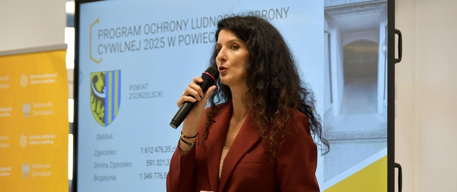 Kobiet z długimi, kręconymi włosami w kolorze czarnym, ubrana w marynarkę w kolorze bordowym stoi na tle ekranu na którym wyświetlana jest prezentacja z rożnym danymi. Kobieta w ręce trzyma mikrofon oraz urządzenie do zmiany slajdów.