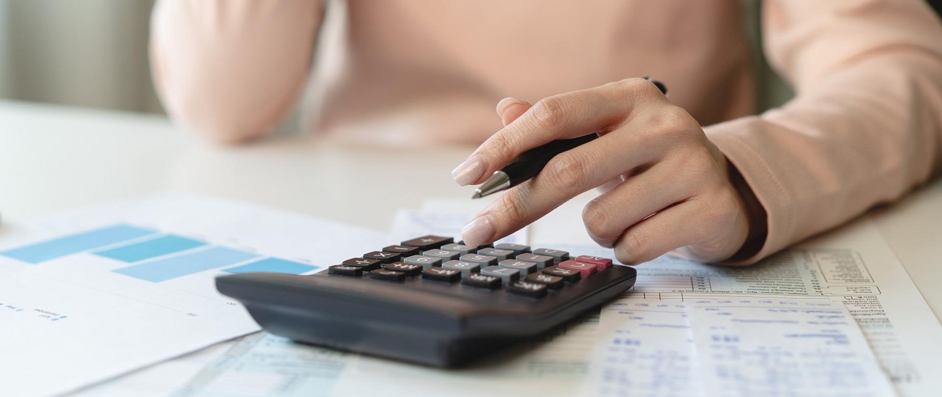 Osoba pracuje na kalkulatorze. Na stoliku leżą faktury
