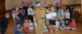 Półkolonia w Śląskowie. W sali wiejskiej uczestnicy półkoloni razem z opiekunami oraz przedstawicielami PSP i Policji pozują do wspólnego zdjęcia na tle mobilnego symulatora zagrożeń pożarowych oraz materiałów promocyjnych. Na pierwszym planie jedna z uczestniczek i zastępca komendanta powiatowego PSP, trzymający dyplom - podziękowanie.