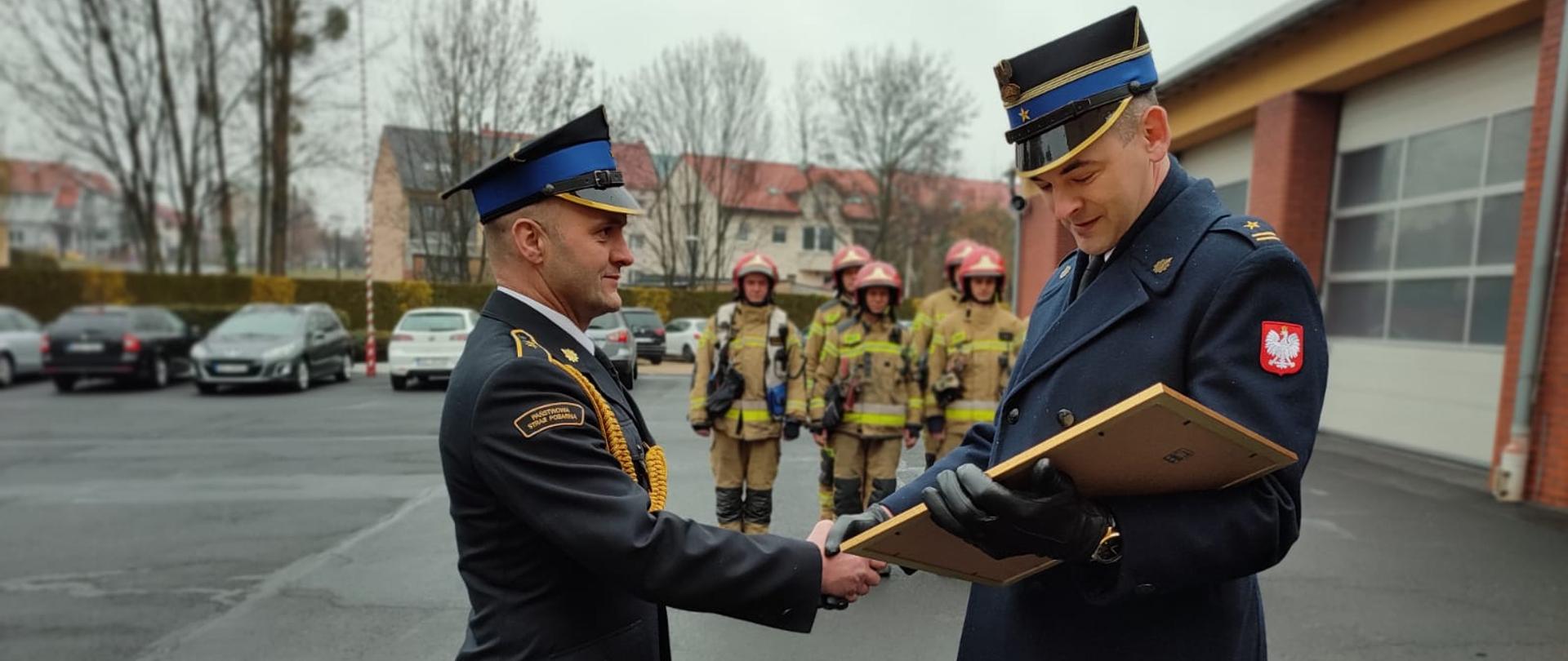 Strażak w płaszczu strażackim wręcza pamiątkową tablicę strażakowi w mundurze wyjściowym. W tle widać strażaków w ubraniach specjalnych koloru beżowego, samochody osobowe, drzewa oraz budynki mieszkalne. 