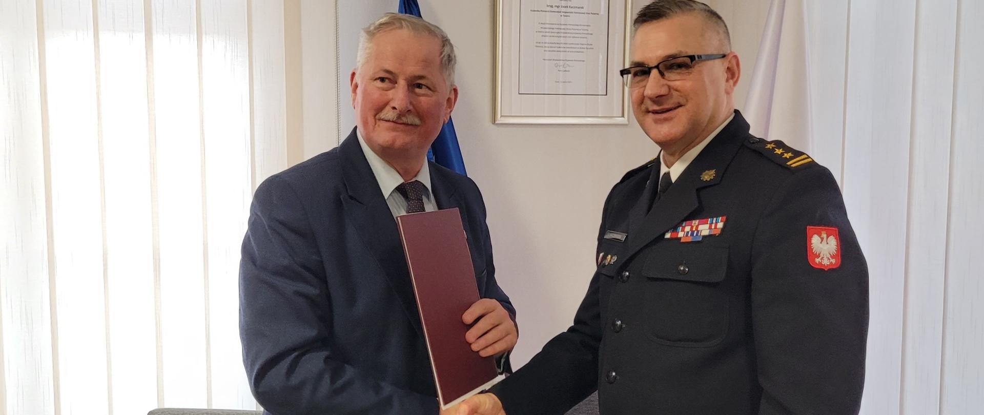 Burmistrz Nowego i komendant wojewódzki PSP stoją przy stole, po podpisaniu umowy i podają sobie dłonie. W ręku Burmistrza podpisany dokument. W tle wystrój gabinetu komendanta.