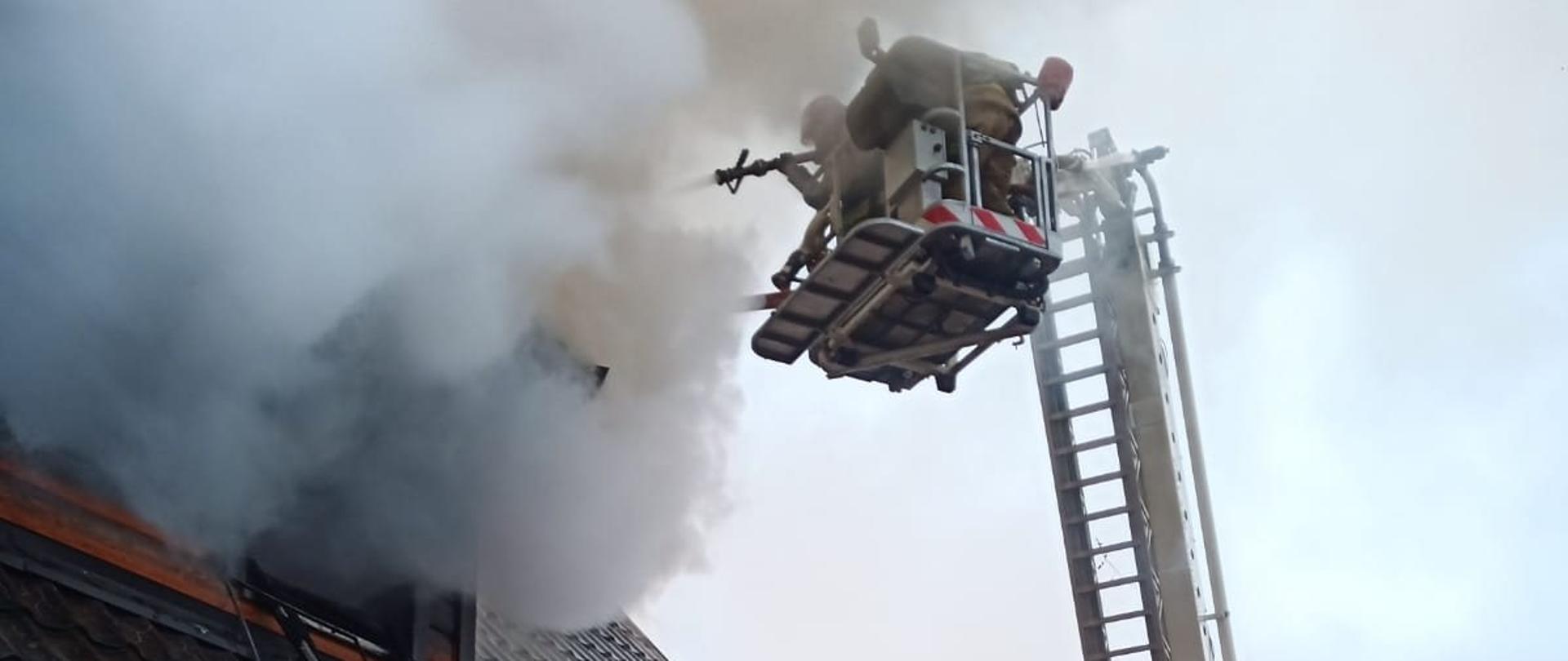 Strażak w koszu podnośnika gasi palący się dach budynku.