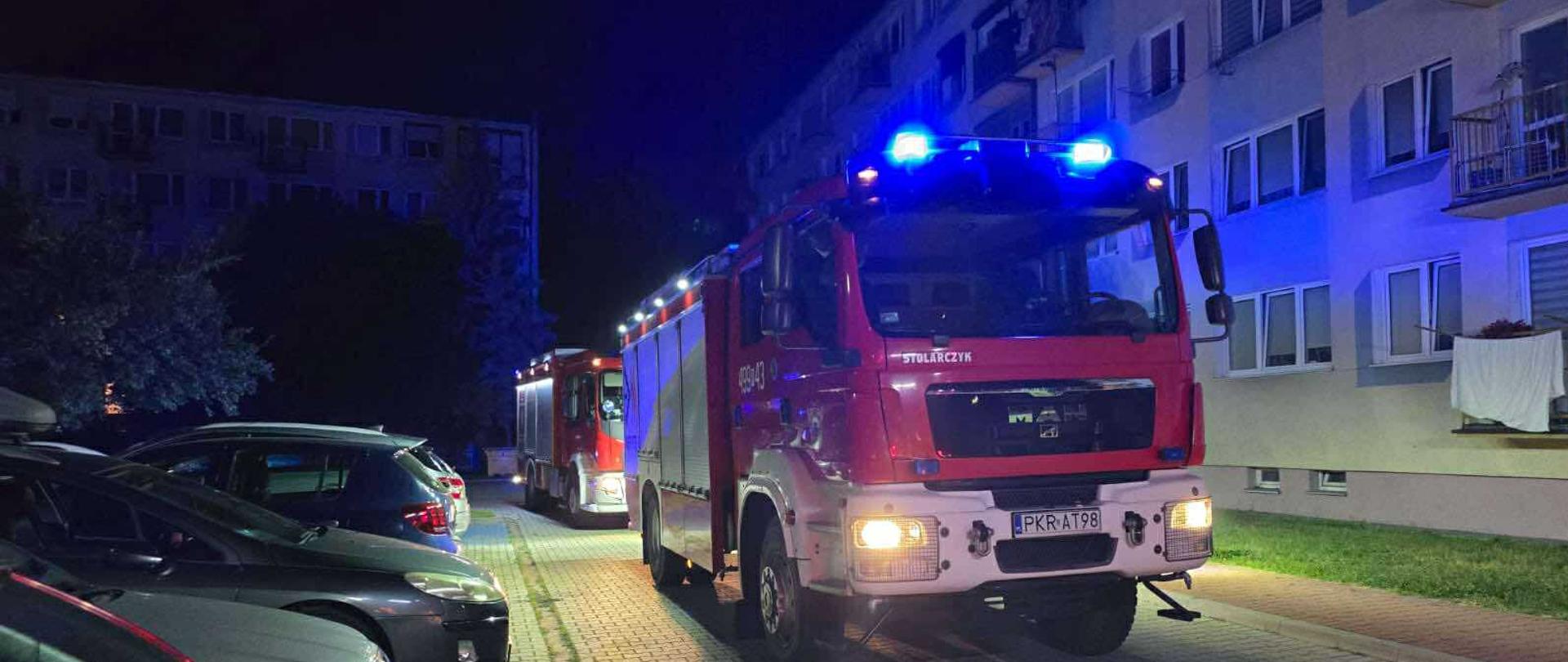Zdjęcie przedstawia pojazdy pożarnicze na drodze dojazdowej do obiektu wielorodzinnego. Na parkingu wzdłuż drogi stoją zaparkowane samochody osobowe