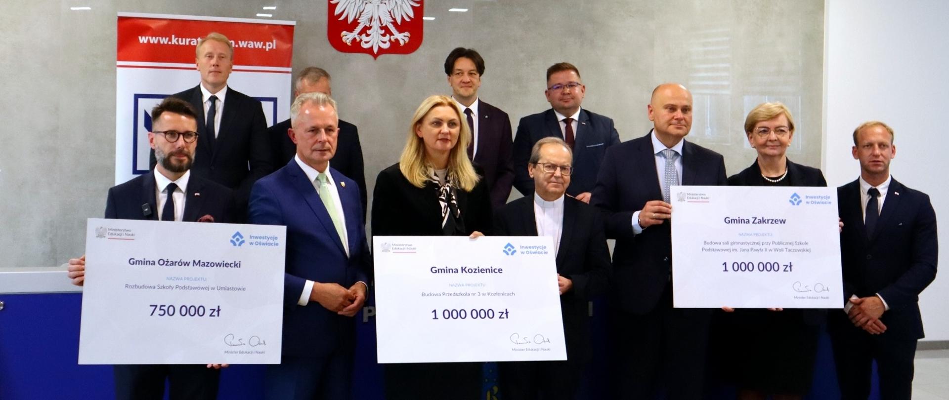 Pod szarą ścianą przed niebieskim stołem stoi grupa dziesięciu osób, trzy osoby z przodu trzymają wielkie symboliczne czeki z napisami Gmina Ożarów Mazowiecki - 750 000 zł, Gmina Kozienice - 1 000 000 zł, Gmina Zakrzew - 1 000 000 zł.