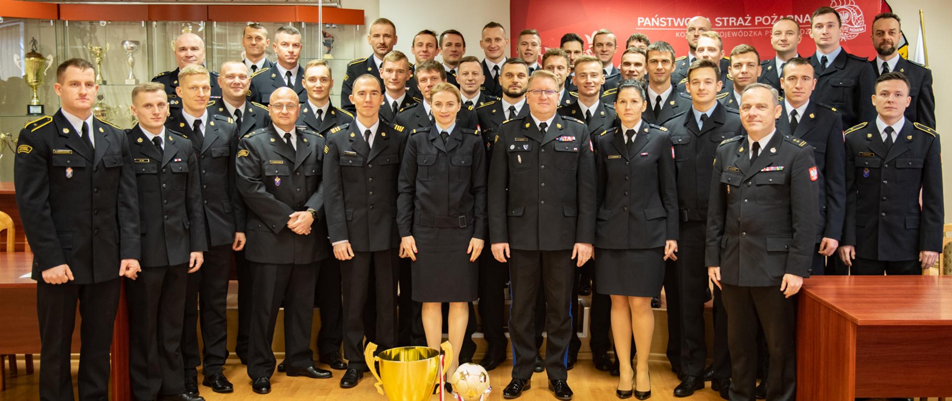 strażacy sportowcy oraz komendant wojewódzki, wszyscy w mundurach wyjściowych pozują do zdjęcia w sali konferencyjnej, w tle za strażakami baner komendy wojewódzkiej oraz przeszklona gablota, przed strażakami stoją dwa puchary