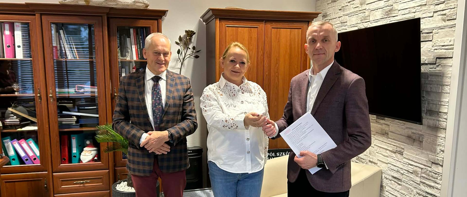 Kobieta wręcza przedmiot mężczyźnie, obok stoi drugi mężczyzna