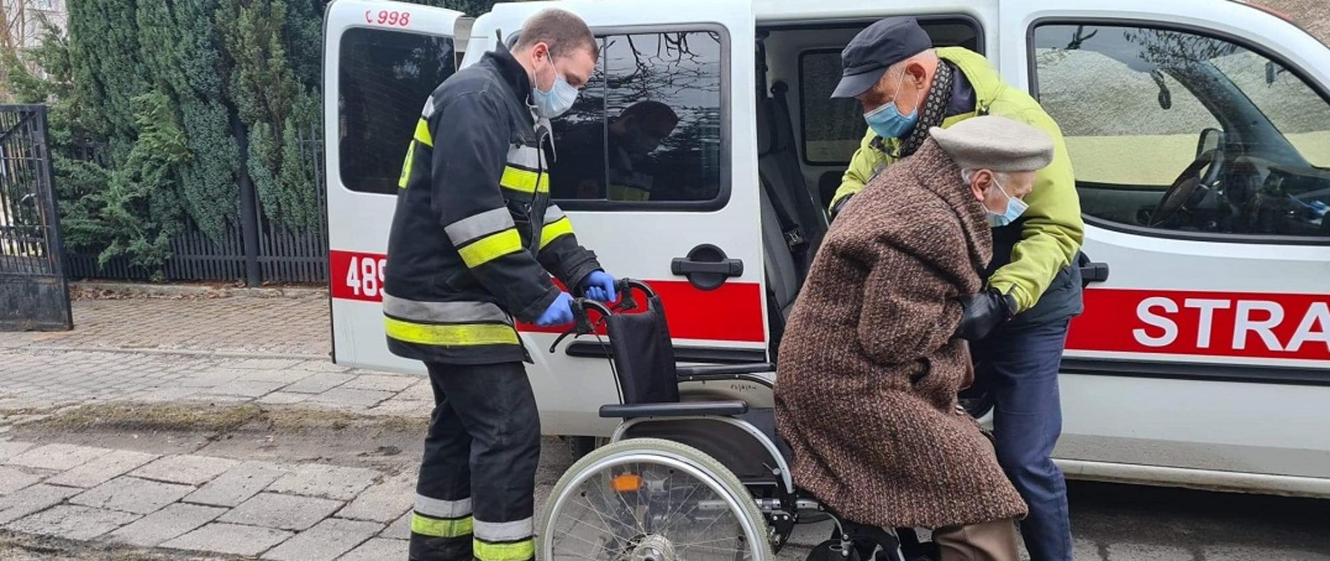 Na zdjęciu widać strażaka oraz mężczyznę pomagających zejść z wózka i wsiąść do samochodu kwatermistrzowskiego Ochotniczej Straży Pożarnej. Kobieta i mężczyzna są spokrewnieni. W tle widać samochód kwatermistrzowski z OSP Skarżysko.