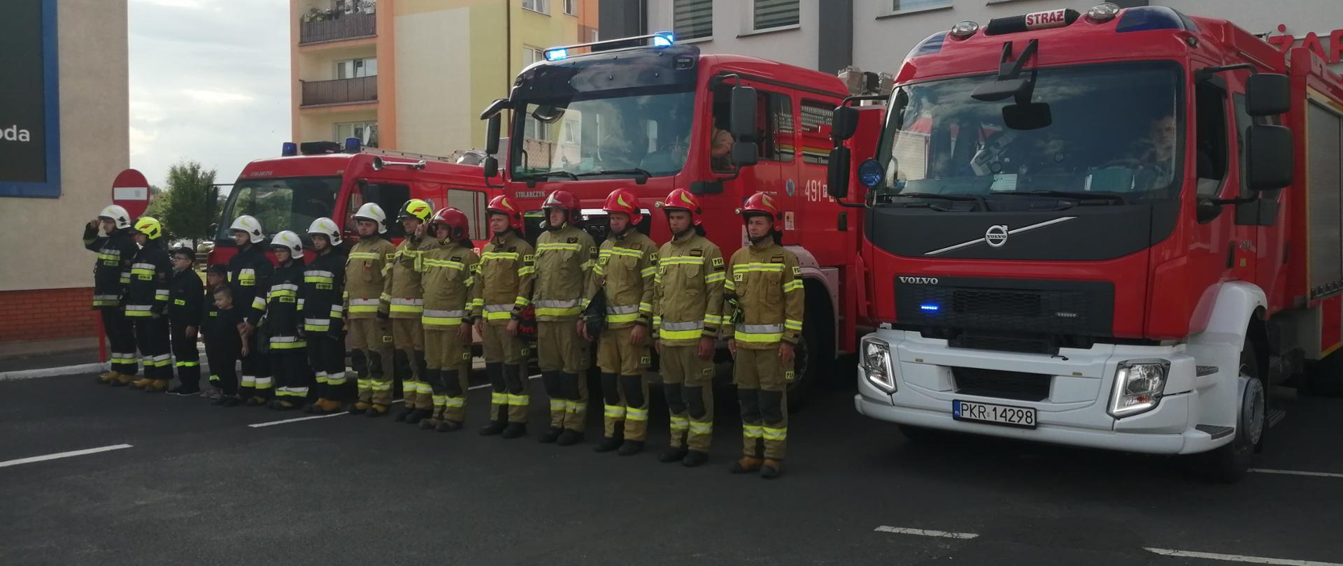 Zdjęcie przedstawia strażaków państwowej i ochotniczej straży pożarnej podczas apelu z okazji Wybuchu Powstania Warszawskiego. Strażacy stoją w mundurach bojowych a za ich plecami samochody pożarnicze.
