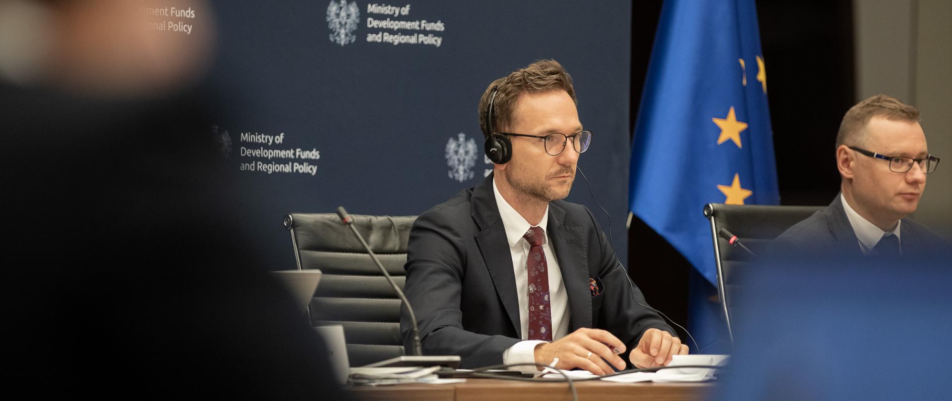 wiceminister Waldemar Buda przy stole uczestniczy w spotkaniu z Komisją Europejską, za nim flaga Unii Europejskiej