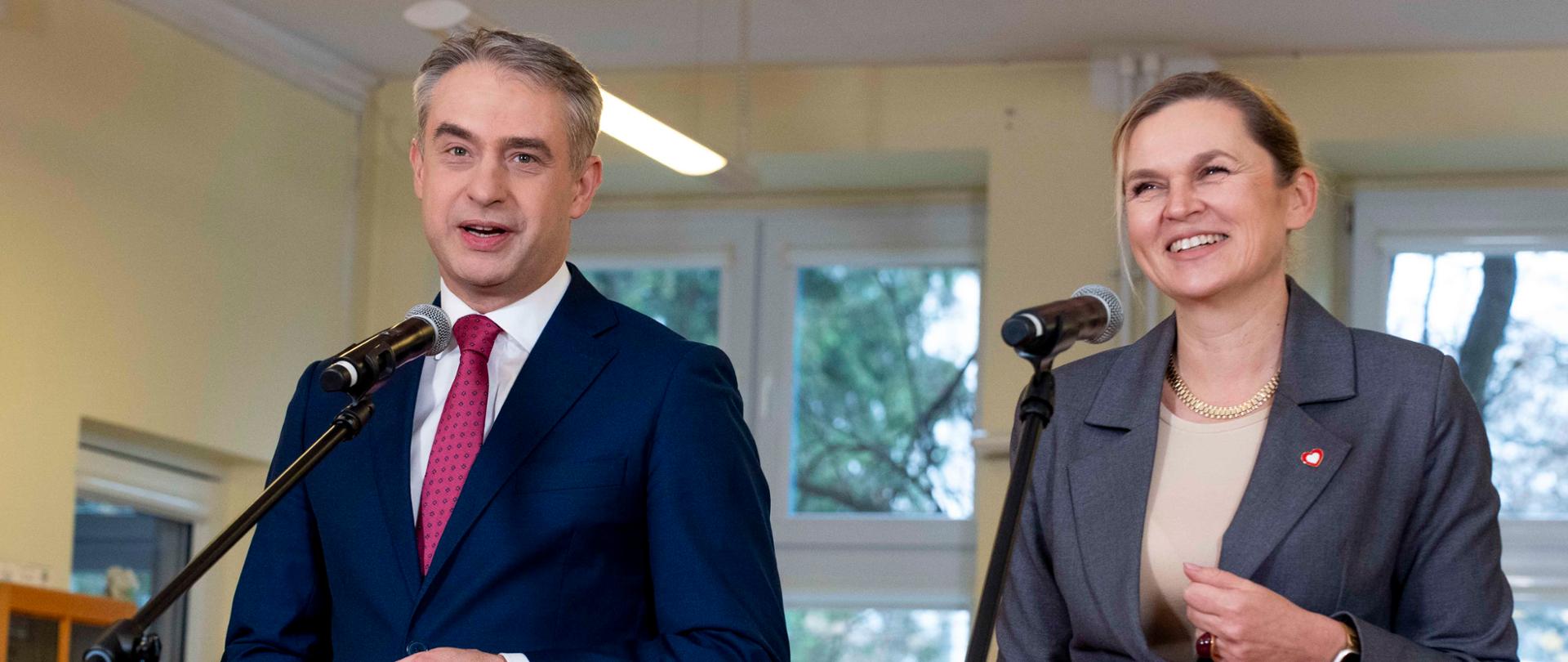 Portretowe ujęcie Krzysztofa Gawkowskiego i Barbary Nowackiej stojących przy mikrofonach. Oboje są uśmiechnięci i patrzą przed siebie. W rozmytym tle widać fragment sali lekcyjnej i uczniów.