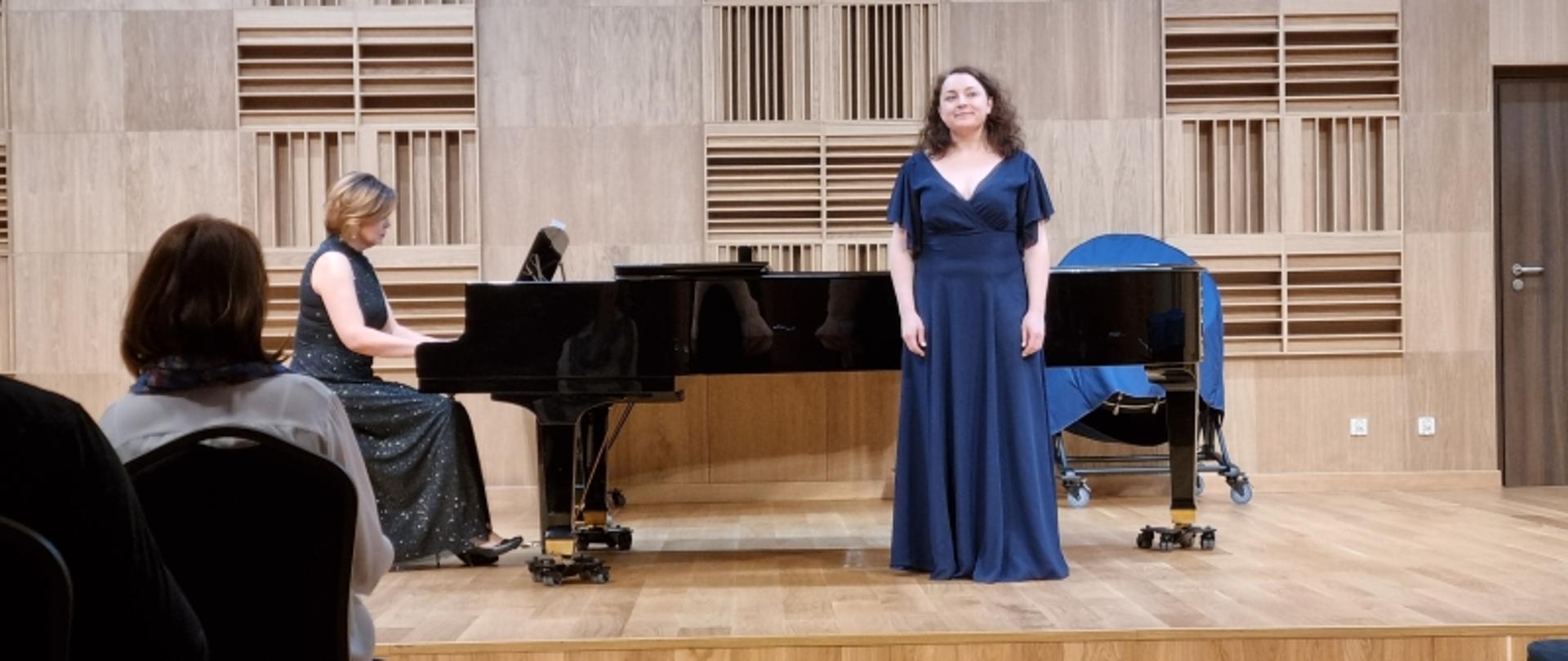 Wokalistka w granatowej sukni z towarzyszeniem pianistki przy fortepianie na estradzie.