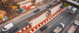 Zdjęcie ukazuje plac budowy wiaduktu nad autostradą. Na pierwszym planie znajdują się dwa filary w trakcie budowy. W tle widoczne są maszyny budowlane, w tym koparki, oraz materiały konstrukcyjne. Pod wiaduktem przebiega autostrada, po której poruszają się samochody osobowe i ciężarówki. Po prawej stronie kadru stoi betonowóz, a po lewej biały van. Cały teren budowy otoczony jest czerwono-białymi barierkami ochronnymi.