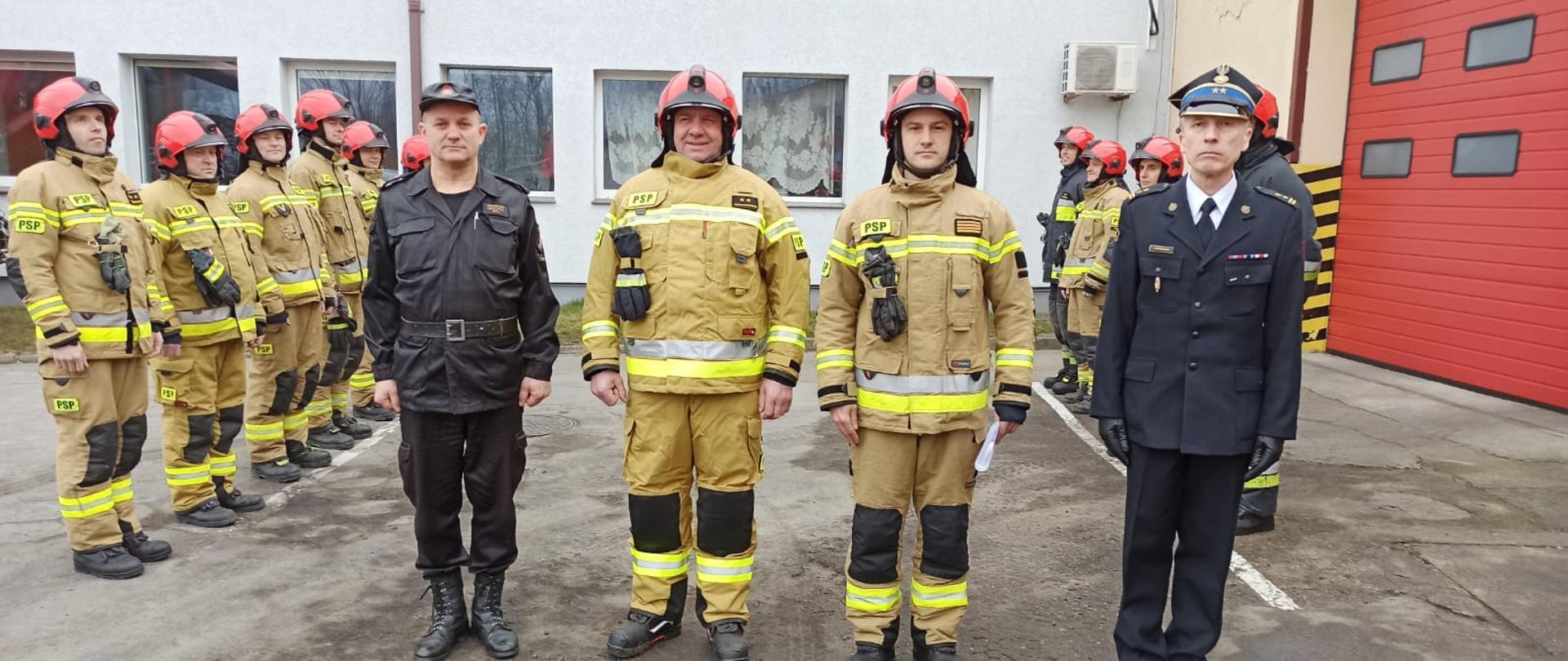 Zdjęcie przedstawia plac przed czerwonymi bramami garażowymi Jednostki Ratowniczo-Gaśniczej w Świdwinie. Przed bramą stoi zmiana służbowa w piaskowych ubraniach specjalnych z czerwonymi hełmami - stoją wzdłuż namalowanej białej linii. Na pierwszym planie znajduje się Komendant Powiatowy PSP w Świdwinie, dowódca JRG ubrani w czarne mundury. Obok nich stoją dwóch wyróżnionych strażaków ubrani w piaskowy mundur i czerwony hełm. W ręku trzymają dokumenty. Panowie się uśmiechają.