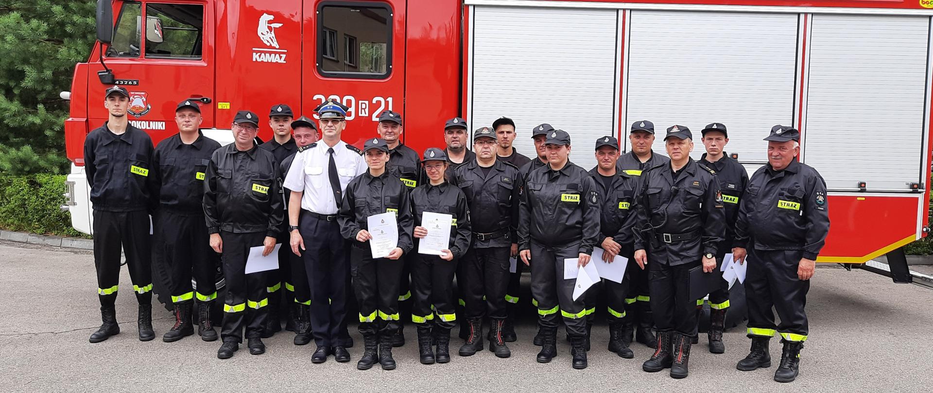 Strażacy w czarnym umundurowaniu stoją przed samochodem ratowniczo-gaśniczym. Jeden ze strażaków występuje w białej koszuli. Niektórzy strażacy trzymają w rękach zaświadczenia o ukończeniu szkolenia podstawowego. 