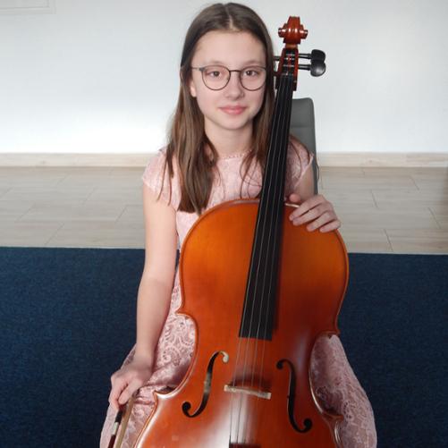 Dziewczynka w okularach siedzi przy wiolonczeli, w ręku trzyma smyczek. Ma na sobie sukienkę w kolorze jasnego różu. W tle biała ściana a na podłodze granatowa wykładzina. 