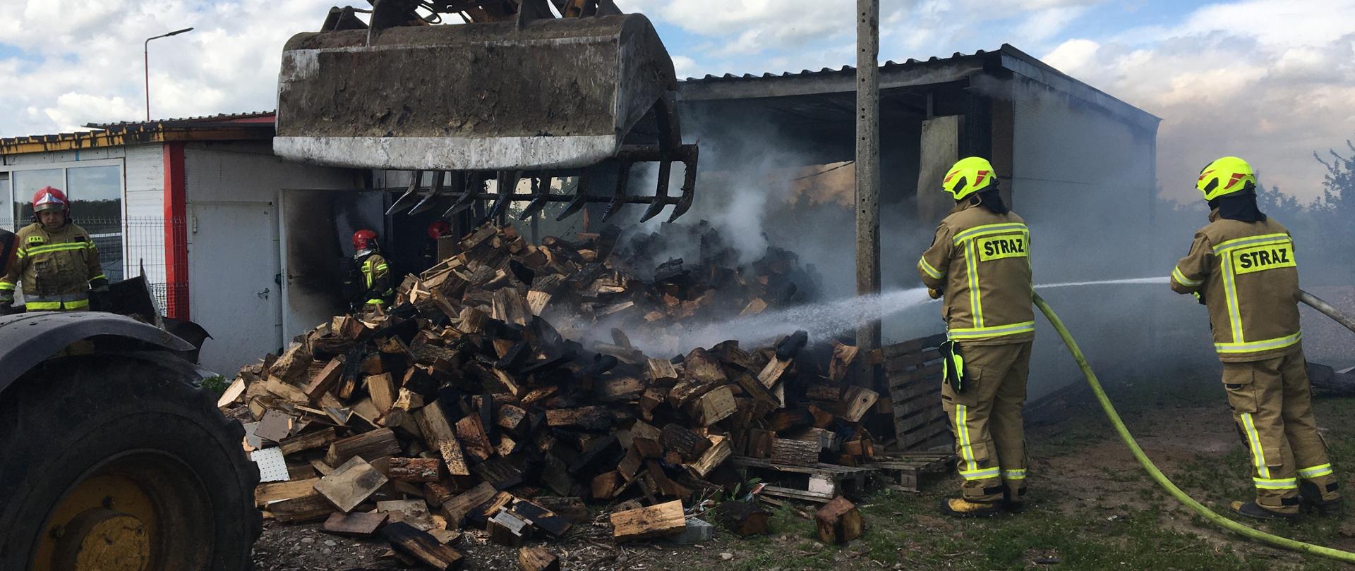 Słoneczny dzień. Sterta drewna opałowego, drewniane palety oraz biała folia przed częściowo spalonym budynkiem. Strażacy w jasnych ubraniach bojowych i żółtych hełmach dogaszają palące się drewno. Przed drewnem koparko-ładowarka rozgarnia stertę drewna.