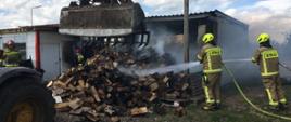 Słoneczny dzień. Sterta drewna opałowego, drewniane palety oraz biała folia przed częściowo spalonym budynkiem. Strażacy w jasnych ubraniach bojowych i żółtych hełmach dogaszają palące się drewno. Przed drewnem koparko-ładowarka rozgarnia stertę drewna