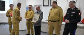 Przedstawiciele Komendy Wojewódzkiej Państwowej Straży Pożarnej wręczają mężczyźnie w garniturze trzymającemu obraz drobny upominek obok stoi kierownictwo Komendy Wojewódzkiej Państwowej Straży Pożarnej. 