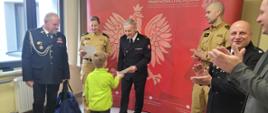 Komendant Miejski w mundurze wyjściowym gratuluje i wręcza dyplom chłoopcu w jaskrawo zielonym polarze. Obok nich inni strażacy w mundurach i pan w marynarce. jeden ze strażaków trzyma w ręce torbę sportową. Za nimi czerwona ścianka firmowa z dużym orłem białym, napisem Państwowa Straż Pożarna i logo PSP.