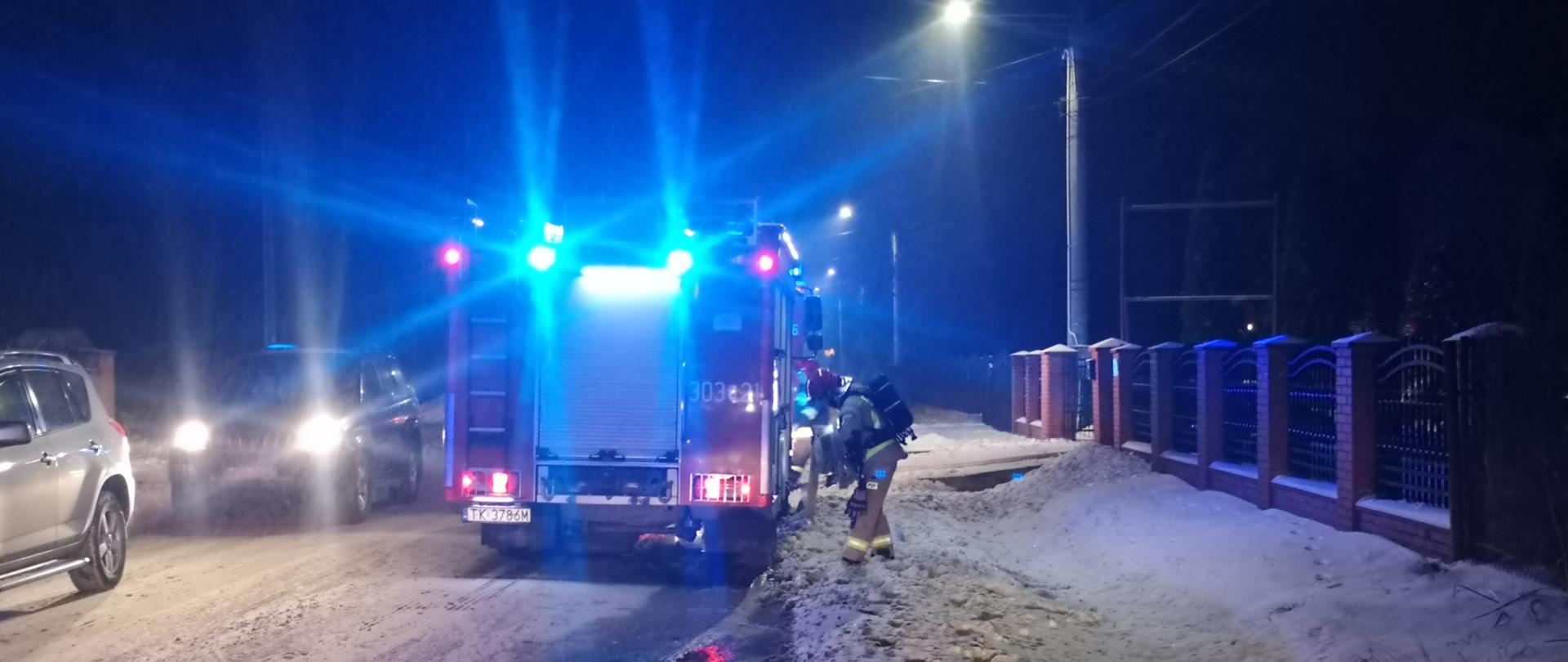 Zdjęcie przedstawia samochód strażacki na sygnałach świetlnych, ustawiony przed posesją na której doszło do pożaru. Wszędzie jest śnieg, na ulicy panuje nie wielki ruch. Przed samochodem znajduje się strażak w aparacie ochrony dróg oddechowych.
