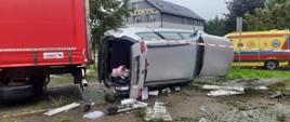 Leżący samochód na boku. Obok stoi karetka Pogotowia oraz widać fragment ciężarówki..