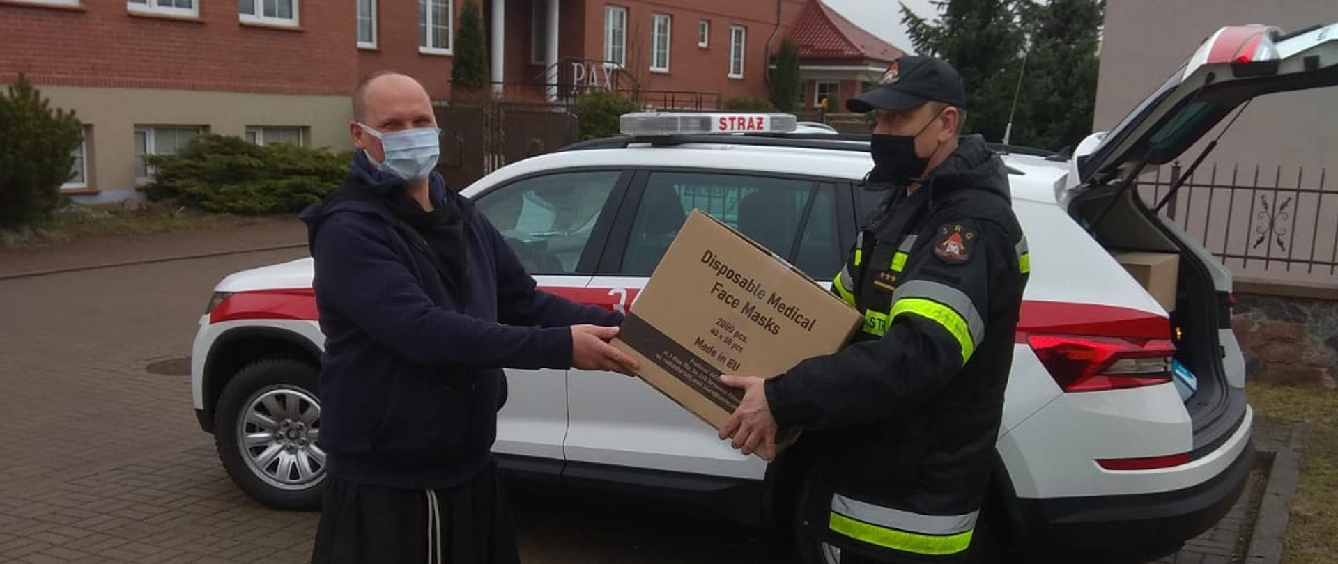 Zdjęcie obrazuje przekazanie księdzu przez strażaka maseczek ochronnych w tle widoczny samochód strażacki i część budynku. 