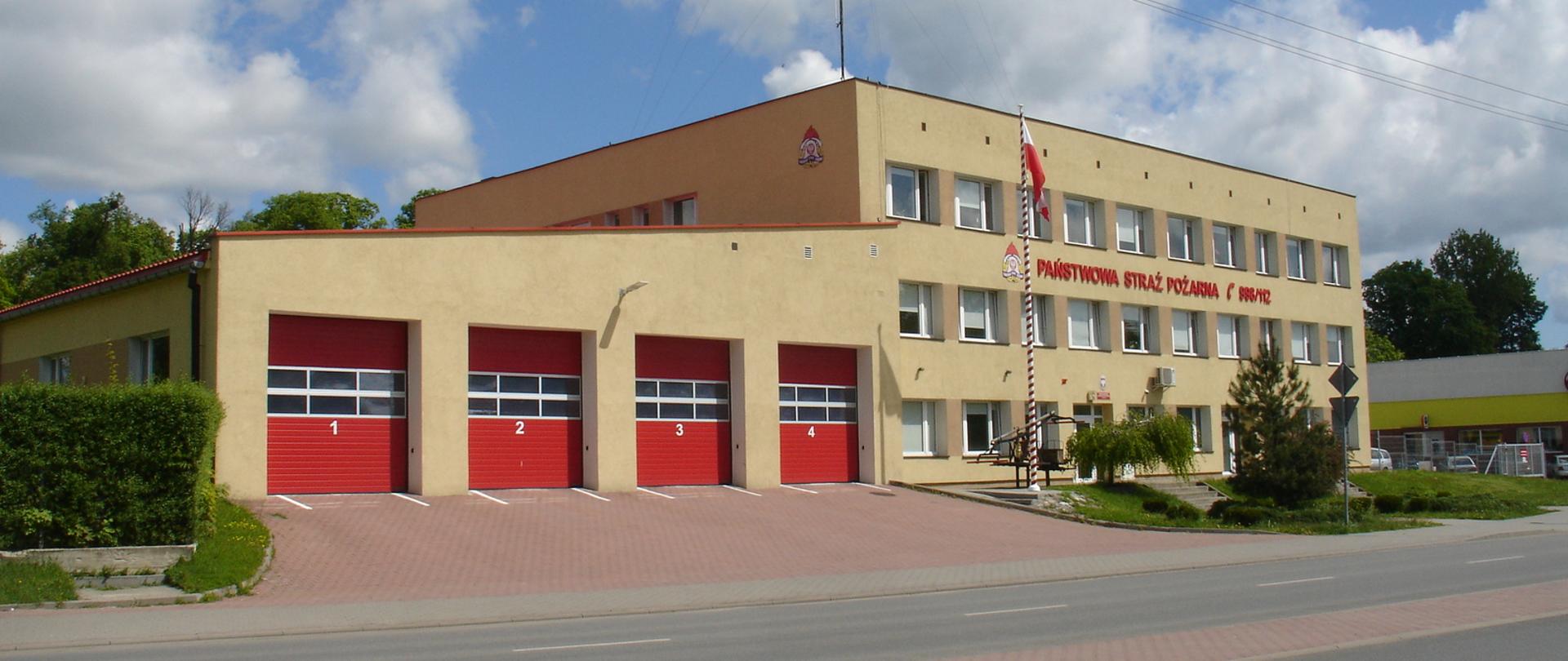 Zdjęcie przedstawia budynek KP PSP w Kętrzynie oraz garaże JRG Kętrzyn. Budynek koloru piaskowego z czerwonymi bramami wyjazdowymi ponumerowanymi od 1 do 4. Na budynku komendy czerwony napis Państwowa Straż Pożarna z logo PSP. Przed budynkiem maszt z wywieszoną biało-czerwoną flagą.