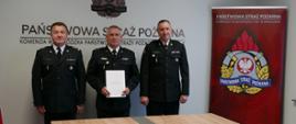 W pomieszczeniu stoją trzej strażacy ubrani w stroje galowe. Po prawej stronie nadbryg. Piotr Filipek Małopolski Komendant Wojewódzki Państwowej Straży Pożarnej, po lewej Komendant Miejski PSP w Tarnowie st. bryg. Krzysztofa Gładysza po środku powołany na stanowisko Zastępcy Komendanta Miejskiego PSP w Tarnowie bryg. Rafała Waśko. Po prawej stronie stojących stażaków znajduje się logo PSP. Przed strażakami znajduje sie długi stół.
