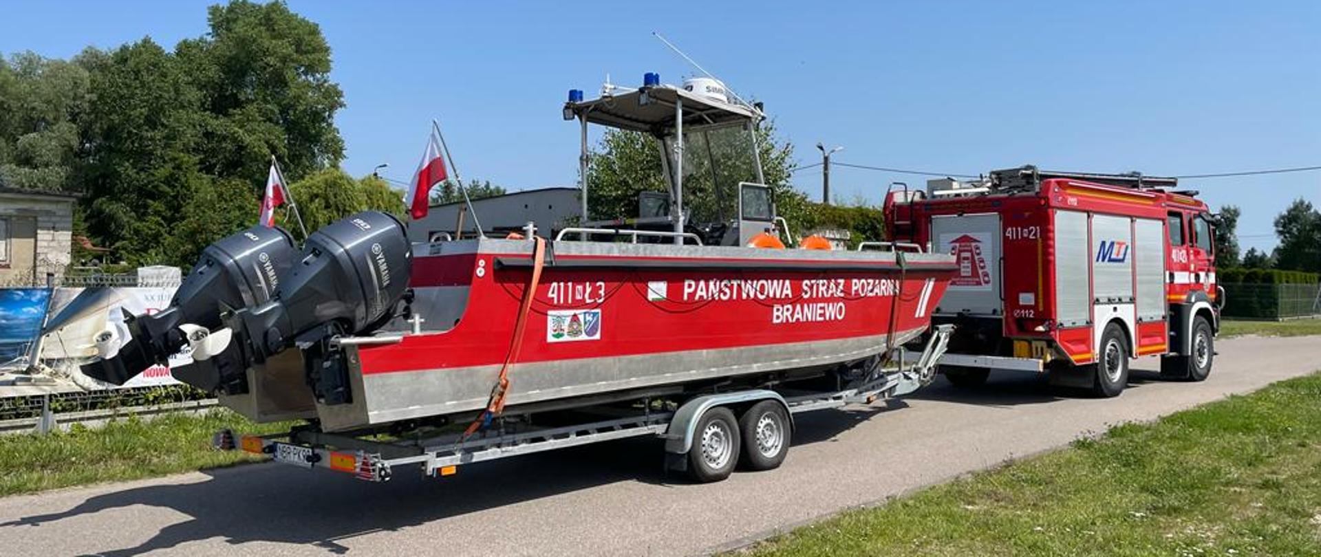 Duża lódź strażacka z dwoma ślinkami na przyczepie podpięta pod samochód strażacki stoi na drodze. Słonecznie.