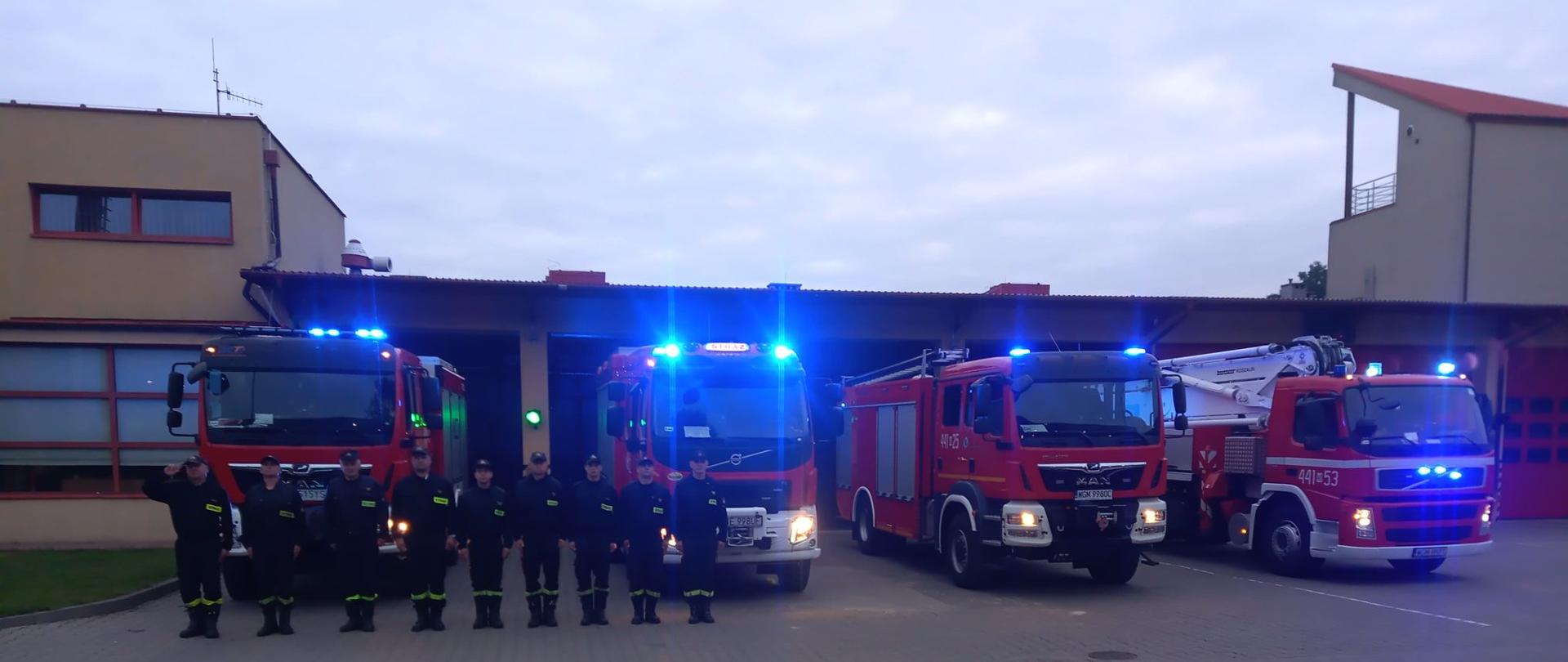 9 strażaków w ubraniach koszarowych stoi przed garażami za nimi stoją samochody ratowniczo-gaśnicze i specjalne z włączonymi sygnałami świetlnymi i dźwiękowymi w celu uczczenia pamięci tragicznie zmarłego strażaka JRG Skierniewice.