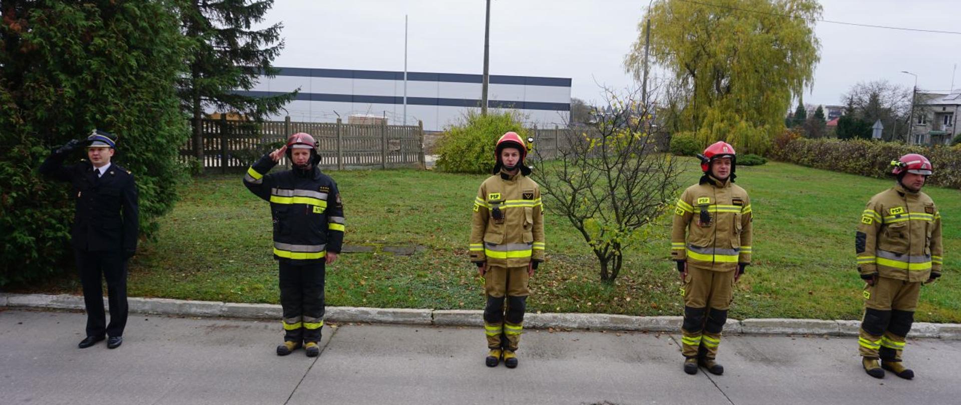 Zdjęcie robione na tle placu Komendy PSP w Hajnówce, widoczna zmiana służbowa wraz z Zastępcą Komendanta Powiatowego PSP w Hajnówce, wszyscy zachowują dystans społeczny, trwa wciąganie flagi państwowej na maszt