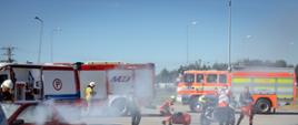 Na zdjęciu przedstawiono moment przemieszczania się osób funkcyjnych i uczestników ćwiczeń w rejonie działań. W pierwszym planie widoczni są przedstawiciele różnych podmiotów zaangażowanych w organizację i przebieg przedsięwzięcia, wyposażeni w kamizelki pozwalające na ich identyfikację.
Fotografia pokazuje również sposób organizacji terenu ćwiczeń oraz obecność pojazdów uczestniczących w działaniach. Istotnym elementem widocznym na zdjęciu jest współpraca osób reprezentujących różne służby i podmioty, która stanowi jeden z kluczowych elementów ćwiczeń ukierunkowanych na sprawdzenie organizacji działań podczas zdarzenia o charakterze masowym.