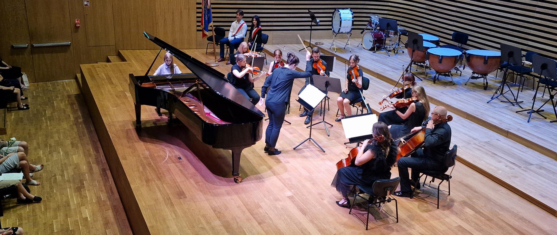 Dziewczynka gra na fortepianie, po prawej stronie dziewięć osób siedzi na krzesłach i gra na skrzypcach, altówkach i wiolonczelach, przed nimi na środku estrady stoi kobieta i dyryguje - ujęcie z góry.