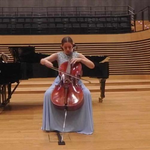 Nastolatka gra na wiolonczeli na środku estrady sali koncertowej PSM, za nią stoi fortepian.