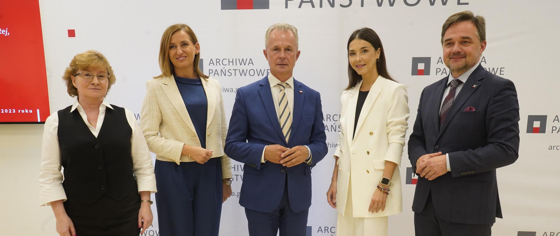 Pod białą ścianą stoi dyrektor Adamiec, mężczyzna w czarnym garniturze i trzy kobiety.