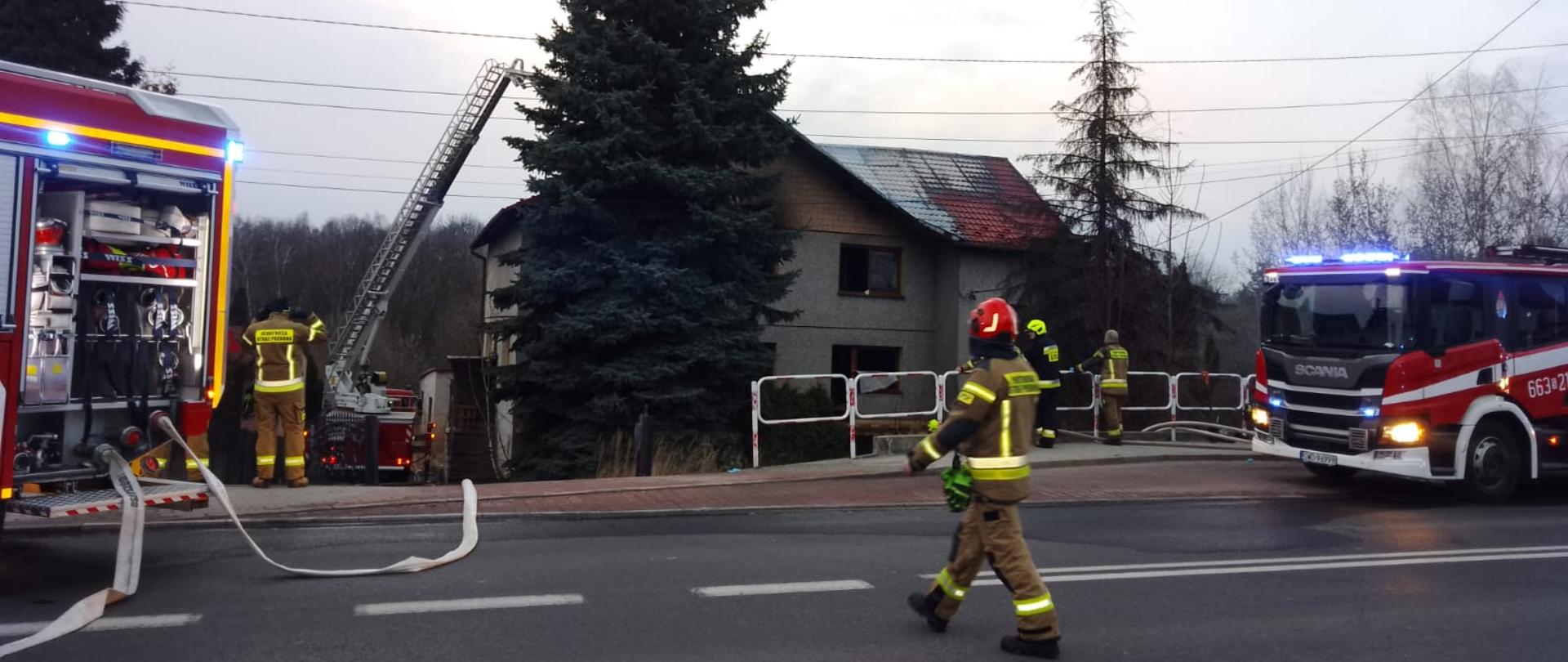 Strażacy prowadzą akcję ratowniczą przy domu jednorodzinnym – na miejscu stoją dwa wozy strażackie z włączonymi sygnałami, a jeden z ratowników przechodzi przez ulicę; do dachu budynku ustawiona jest drabina.